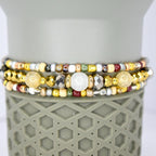 Three beaded bracelets with a variety of colors and textures. The bracelets feature gold, silver, and bronze beads, along with larger, round, and faceted beads. The bracelets are stacked on top of a gray, textured surface.
