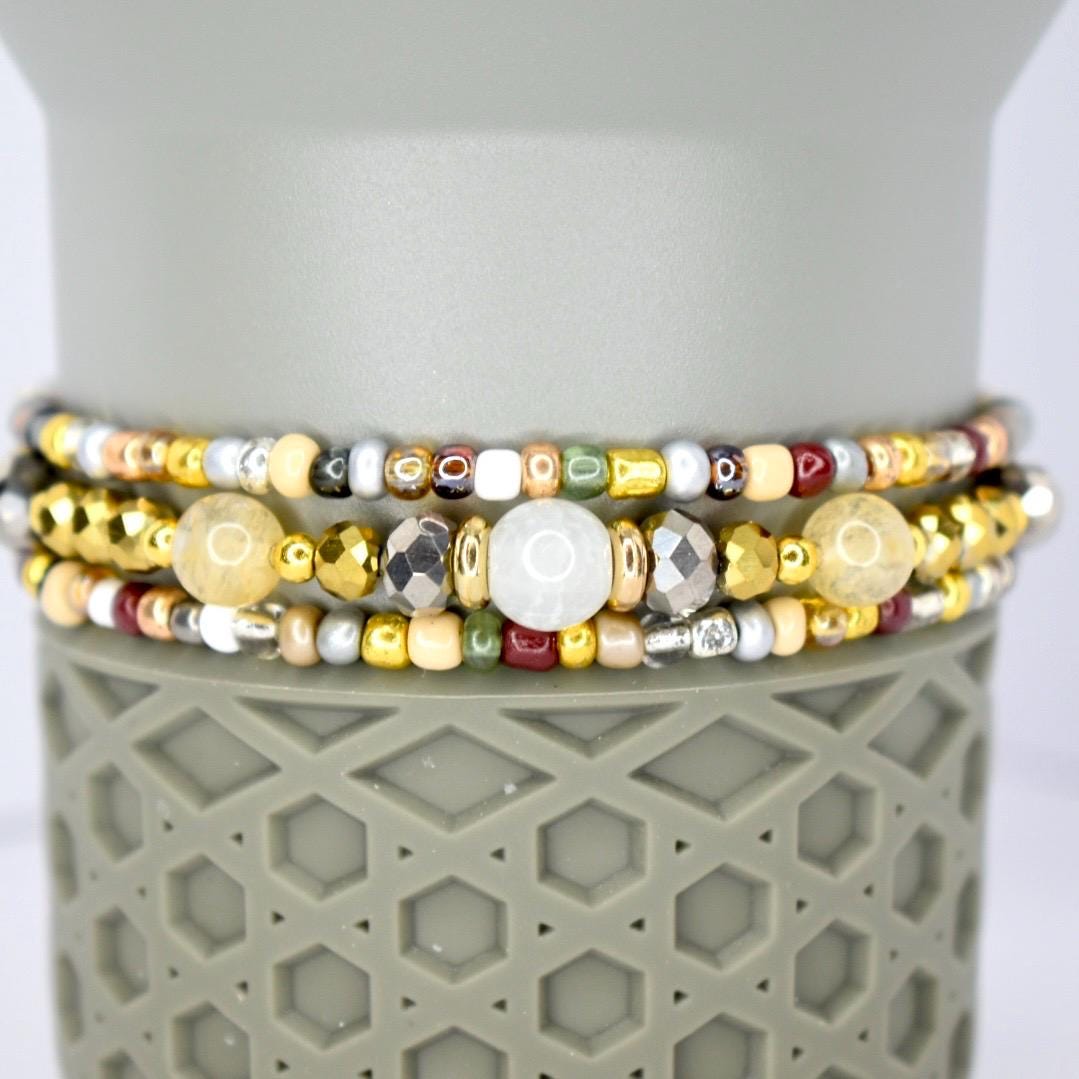 Three beaded bracelets with a variety of colors and textures. The bracelets feature gold, silver, and bronze beads, along with larger, round, and faceted beads. The bracelets are stacked on top of a gray, textured surface.