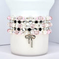 Stacked beaded bracelets in pink, white, black, and silver. The bracelets feature pearls, faceted beads, and small silver accents. A silver bow charm hangs from the bottom bracelet. The bracelets are displayed on a white cylindrical object.