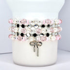 Stacked beaded bracelets in pink, white, black, and silver. The bracelets feature pearls, faceted beads, and small silver accents. A silver bow charm hangs from the bottom bracelet. The bracelets are displayed on a white cylindrical object.