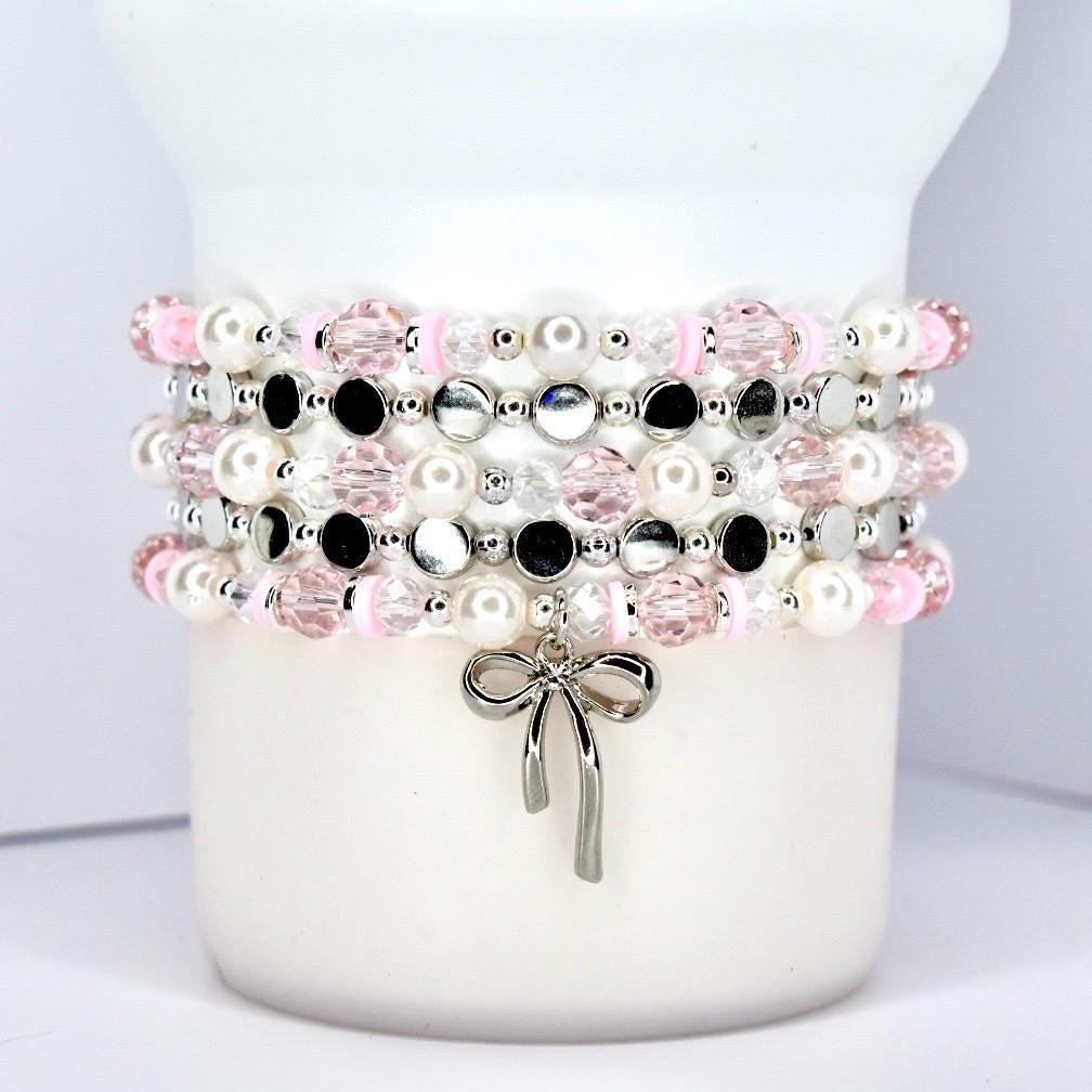 Stacked beaded bracelets in pink, white, black, and silver. The bracelets feature pearls, faceted beads, and small silver accents. A silver bow charm hangs from the bottom bracelet. The bracelets are displayed on a white cylindrical object.