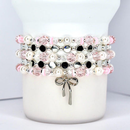 Stacked beaded bracelets in pink, white, black, and silver. The bracelets feature pearls, faceted beads, and small silver accents. A silver bow charm hangs from the bottom bracelet. The bracelets are displayed on a white cylindrical object.