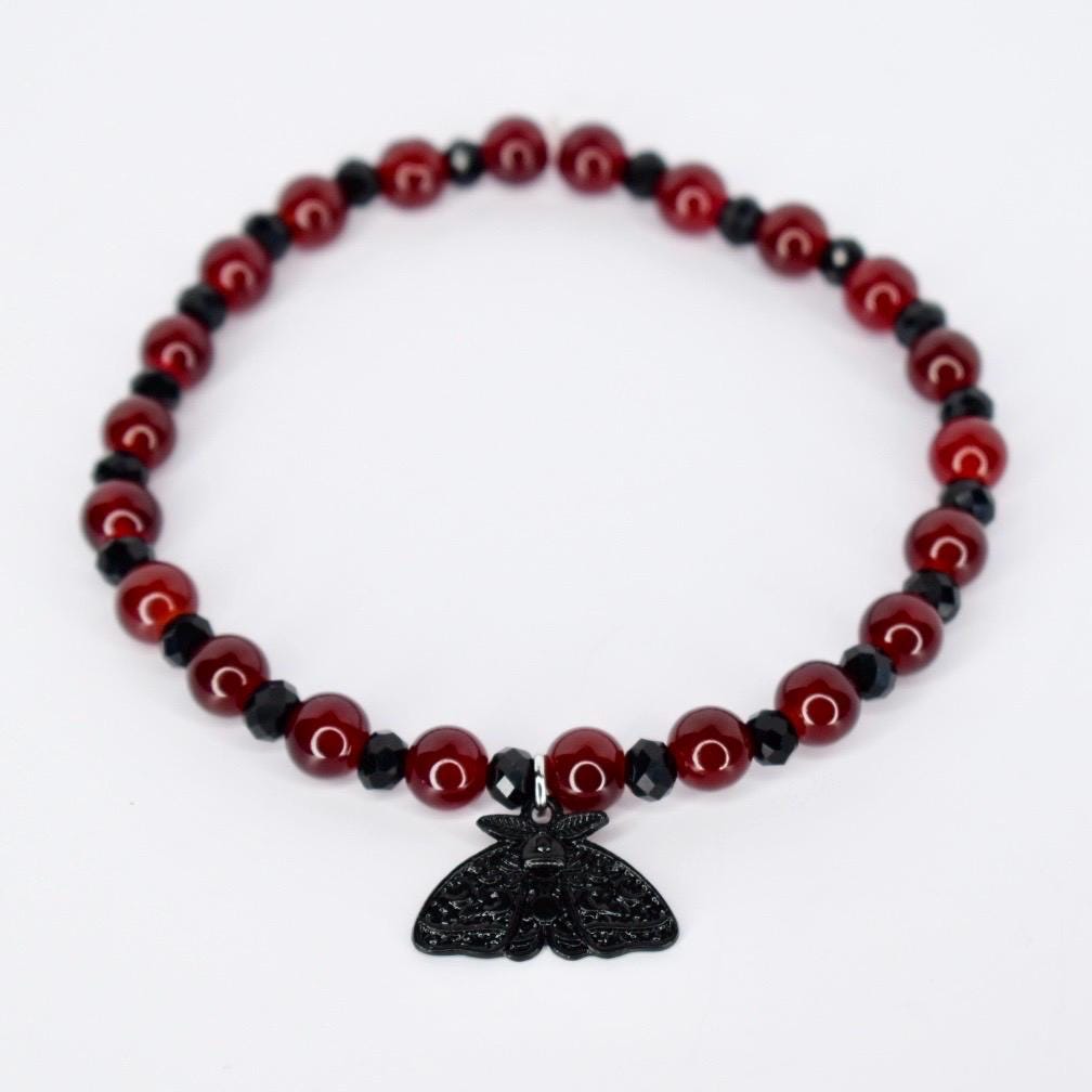 A beaded bracelet featuring alternating red and black beads. The red beads are round and glossy, while the black beads are faceted. A black moth pendant hangs from the center of the bracelet, adding a gothic or alternative aesthetic. The bracelet is likely elasticized for easy wear.
