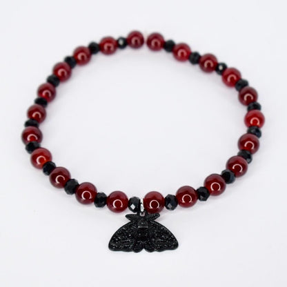 A beaded bracelet featuring alternating red and black beads. The red beads are round and glossy, while the black beads are faceted. A black moth pendant hangs from the center of the bracelet, adding a gothic or alternative aesthetic. The bracelet is likely elasticized for easy wear.
