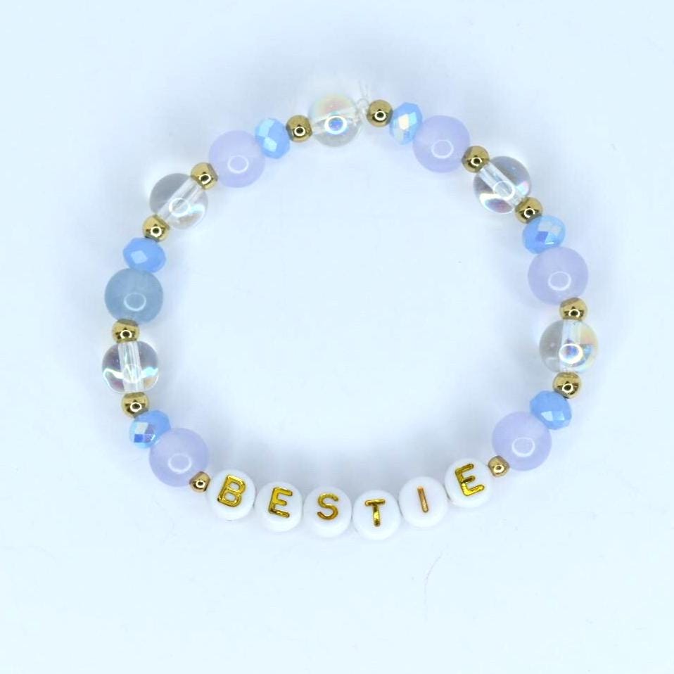 A beaded bracelet with the word 'BESTIE' spelled out in gold letters on white beads. The bracelet features a combination of light blue, clear, and gold-colored beads. The beads are arranged in a circular pattern, creating a stylish accessory.
