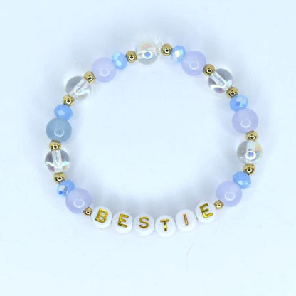 A beaded bracelet with the word 'BESTIE' spelled out in gold letters on white beads. The bracelet features a combination of light blue, clear, and gold-colored beads. The beads are arranged in a circular pattern, creating a stylish accessory.