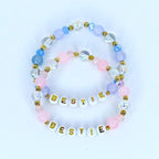 Two beaded bracelets with the word 'BESTIE' spelled out in gold letters. The bracelets feature a combination of white, pink, light blue, and clear beads. The beads are arranged in a pattern with gold accents, creating a friendship-themed accessory.