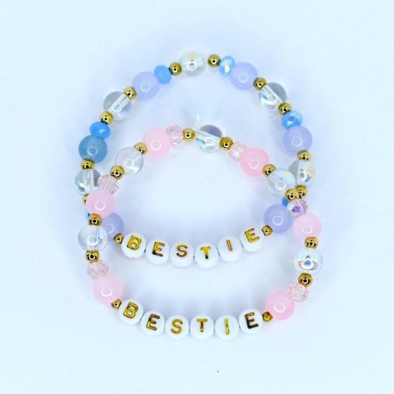 Two beaded bracelets with the word 'BESTIE' spelled out in gold letters. The bracelets feature a combination of white, pink, light blue, and clear beads. The beads are arranged in a pattern with gold accents, creating a friendship-themed accessory.