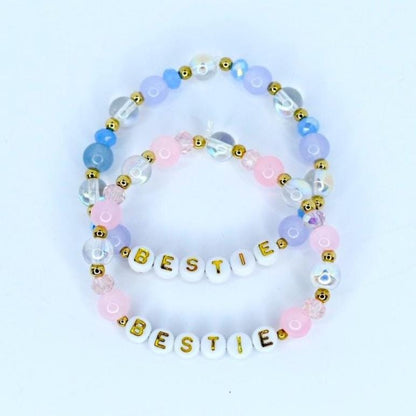 Two beaded bracelets with the word 'BESTIE' spelled out in gold letters. The bracelets feature a combination of white, pink, light blue, and clear beads. The beads are arranged in a pattern with gold accents, creating a friendship-themed accessory.
