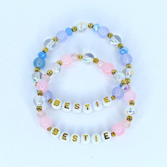 Two beaded bracelets with the word 'BESTIE' spelled out in gold letters. The bracelets feature a combination of white, pink, light blue, and clear beads. The beads are arranged in a pattern with gold accents, creating a friendship-themed accessory.