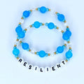 Two beaded bracelets. The top bracelet features light blue and clear beads with gold accents. The bottom bracelet has the word "RESILIENT" spelled out in white letter beads. The bracelets are on a white background.