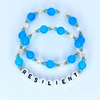 Two beaded bracelets. The top bracelet features light blue and clear beads with gold accents. The bottom bracelet has the word "RESILIENT" spelled out in white letter beads. The bracelets are on a white background.