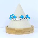Two beaded bracelets with the word "RESILIENT" spelled out in white letter beads. The bracelets feature blue and clear beads, with gold accents. The bracelets are displayed on a white cone-shaped stand with a wooden base.