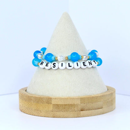Two beaded bracelets with the word "RESILIENT" spelled out in white letter beads. The bracelets feature blue and clear beads, with gold accents. The bracelets are displayed on a white cone-shaped stand with a wooden base.