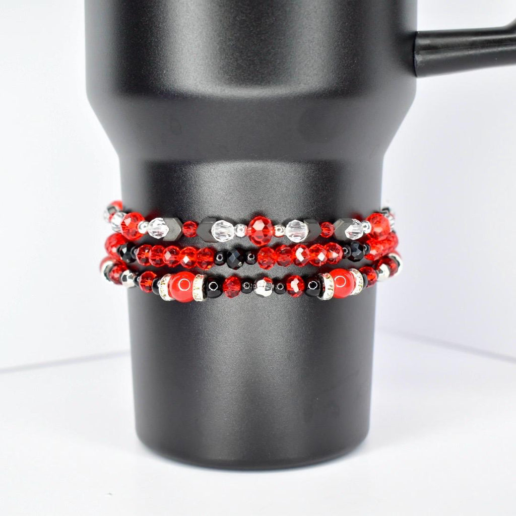 Three beaded bracelets in red, black, and silver are stacked on a black tumbler. The top bracelet features red and clear beads with black accents. The middle bracelet has red, black, and silver beads. The bottom bracelet has large red beads with black and silver accents.