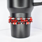 Three beaded bracelets in red, black, and silver are stacked on a black tumbler. The top bracelet features red and clear beads with black accents. The middle bracelet has red, black, and silver beads. The bottom bracelet has large red beads with black and silver accents.