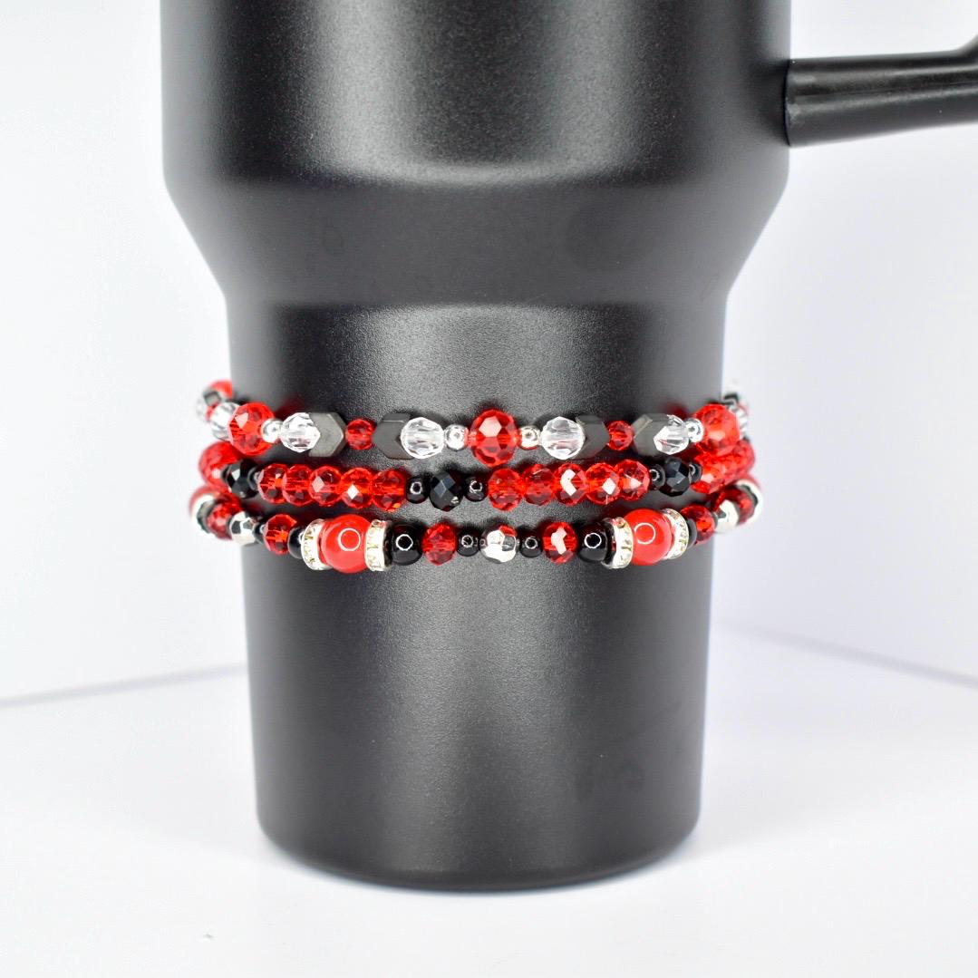 Three beaded bracelets in red, black, and silver are stacked on a black tumbler. The top bracelet features red and clear beads with black accents. The middle bracelet has red, black, and silver beads. The bottom bracelet has large red beads with black and silver accents.