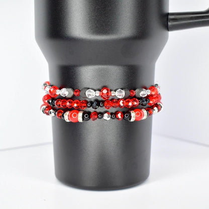 Three beaded bracelets in red, black, and silver are stacked on a black tumbler. The top bracelet features red and clear beads with black accents. The middle bracelet has red, black, and silver beads. The bottom bracelet has large red beads with black and silver accents.