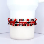 Three beaded bracelets in red, black, and silver. The top bracelet has red and clear beads with black arrow-shaped beads. The middle bracelet features red and black beads. The bottom bracelet has red and black beads with silver accents. The bracelets are stacked on a white cylindrical surface.