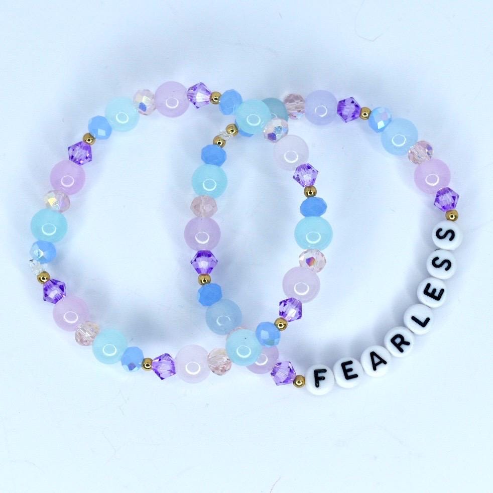 Two beaded bracelets with a pastel color scheme. One bracelet features light blue, pink, and purple beads with gold accents. The other bracelet has the word 'FEARLESS' spelled out in white letter beads. The bracelets are a fashionable accessory.
