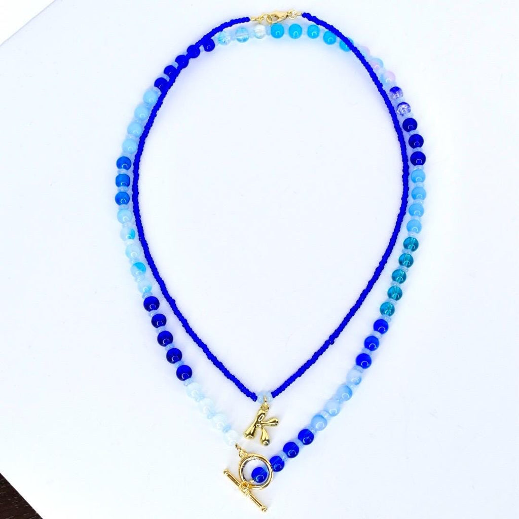 Two layered beaded necklaces in shades of blue. The top necklace features small blue beads, while the bottom one has larger beads and a gold-colored charm with the letter 'K'. Both necklaces have a gold-colored clasp.