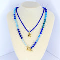 Two layered necklaces displayed on a mannequin. The top necklace is a thin, dark blue beaded chain with a gold letter 'K' pendant. The bottom necklace is a thicker chain with blue and light blue beads, and a gold toggle clasp.