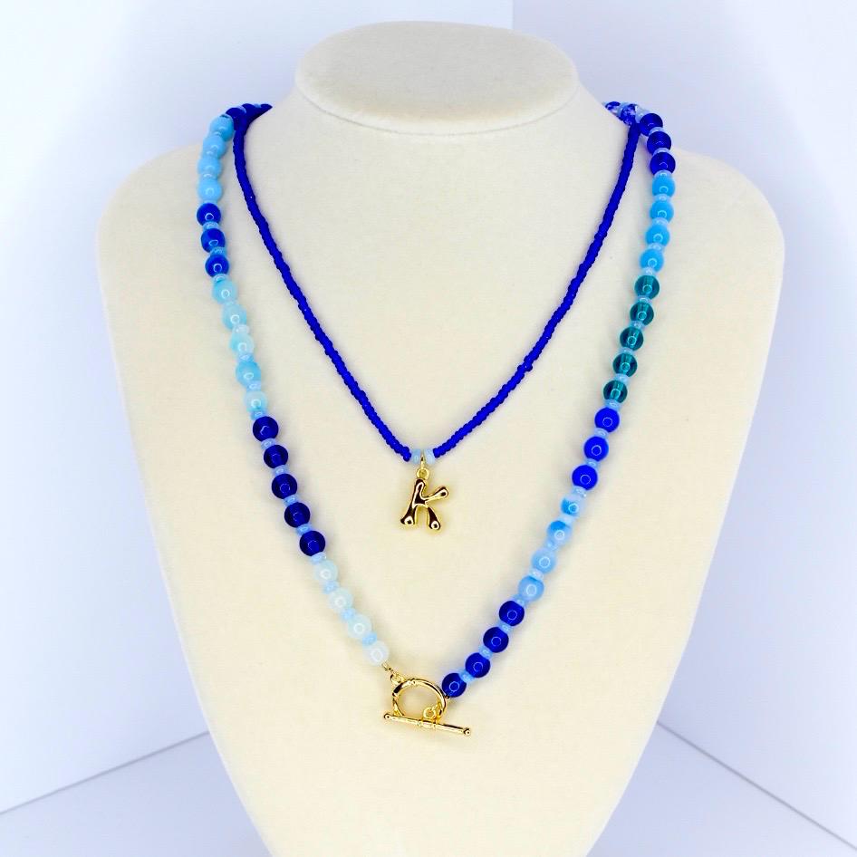 Two layered necklaces displayed on a mannequin. The top necklace is a thin, dark blue beaded chain with a gold letter 'K' pendant. The bottom necklace is a thicker chain with blue and light blue beads, and a gold toggle clasp.