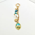 A gold-colored keychain with a lobster clasp and a chain holding several charms. The charms include a large white pearl, a seashell, and a blue and gold oval pendant with a floral design. Other charms include a small pearl and a blue leaf.