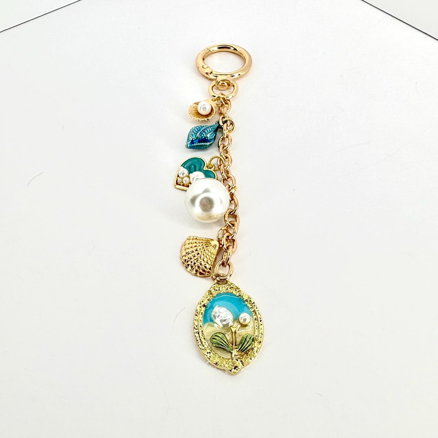 A gold-colored keychain with a lobster clasp and a chain holding several charms. The charms include a large white pearl, a seashell, and a blue and gold oval pendant with a floral design. Other charms include a small pearl and a blue leaf.