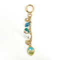 Gold-tone keychain with a lobster clasp, featuring a chain and several charms. Charms include a seashell, a blue leaf, a teal heart with pearls, a large white pearl, a seashell, and an oval pendant with a floral design.