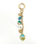 Gold-tone keychain with a lobster clasp, featuring a chain and several charms. Charms include a seashell, a blue leaf, a teal heart with pearls, a large white pearl, a seashell, and an oval pendant with a floral design.