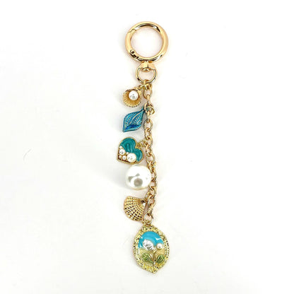 Gold-tone keychain with a lobster clasp, featuring a chain and several charms. Charms include a seashell, a blue leaf, a teal heart with pearls, a large white pearl, a seashell, and an oval pendant with a floral design.