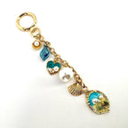 Gold-tone keychain with a variety of ocean-themed charms. The charms include a blue leaf, a teal heart with pearls, a large white pearl, a seashell, and an oval charm with a seashell design. The keychain is a decorative accessory.