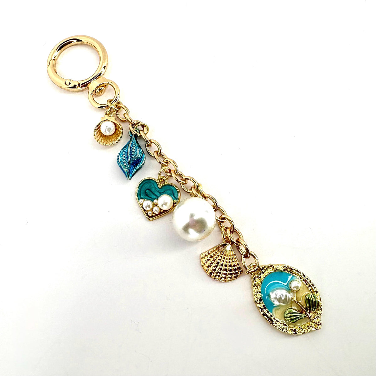 Gold-tone keychain with a variety of ocean-themed charms. The charms include a blue leaf, a teal heart with pearls, a large white pearl, a seashell, and an oval charm with a seashell design. The keychain is a decorative accessory.