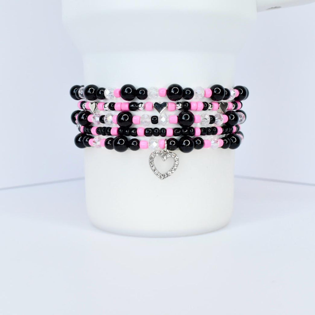 Stack of beaded bracelets in black, pink, and clear beads. Each bracelet has a different pattern, with a silver heart charm dangling from the bottom bracelet. The bracelets are displayed on a white cylindrical object.