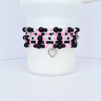 Stack of beaded bracelets in black, pink, and clear beads. Each bracelet has a different pattern, with a silver heart charm dangling from the bottom bracelet. The bracelets are displayed on a white cylindrical object.