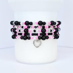 Stack of beaded bracelets in black, pink, and clear beads. Each bracelet has a different design, with a silver heart charm dangling from the bottom bracelet. The bracelets are displayed on a white surface.