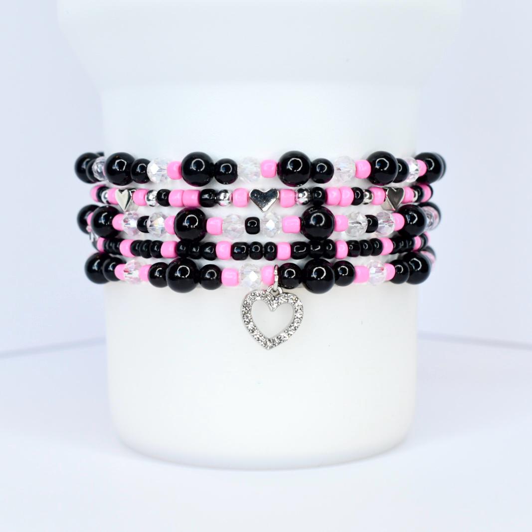 Stack of beaded bracelets in black, pink, and clear beads. Each bracelet has a different design, with a silver heart charm dangling from the bottom bracelet. The bracelets are displayed on a white surface.