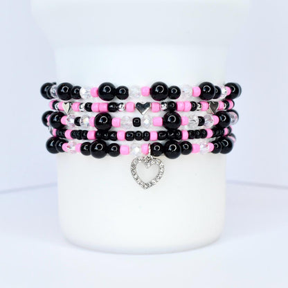 Stack of beaded bracelets in black, pink, and clear beads. Each bracelet has a different design, with a silver heart charm dangling from the bottom bracelet. The bracelets are displayed on a white surface.