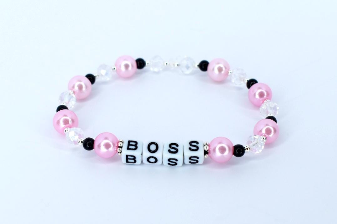 A pink and white beaded bracelet with the word 'BOSS' spelled out in white letter beads. The bracelet features pink pearl beads, clear crystal beads, and small black beads. The bracelet is displayed on a white surface.