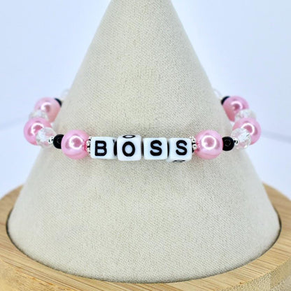 A pink and white beaded bracelet spelling out the word "BOSS". The bracelet features pink pearl beads, clear crystal beads, and black accent beads. The letters are white with black lettering.