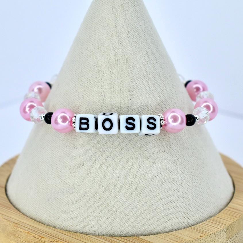 A pink and white beaded bracelet spelling out the word "BOSS". The bracelet features pink pearl beads, clear crystal beads, and black accent beads. The letters are white with black lettering.