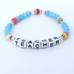 A beaded bracelet with the word "TEACHER" spelled out in white letter beads. The bracelet features light blue beads, silver accents, and colorful accent beads in pink, yellow, orange, and blue. A perfect gift for a teacher.