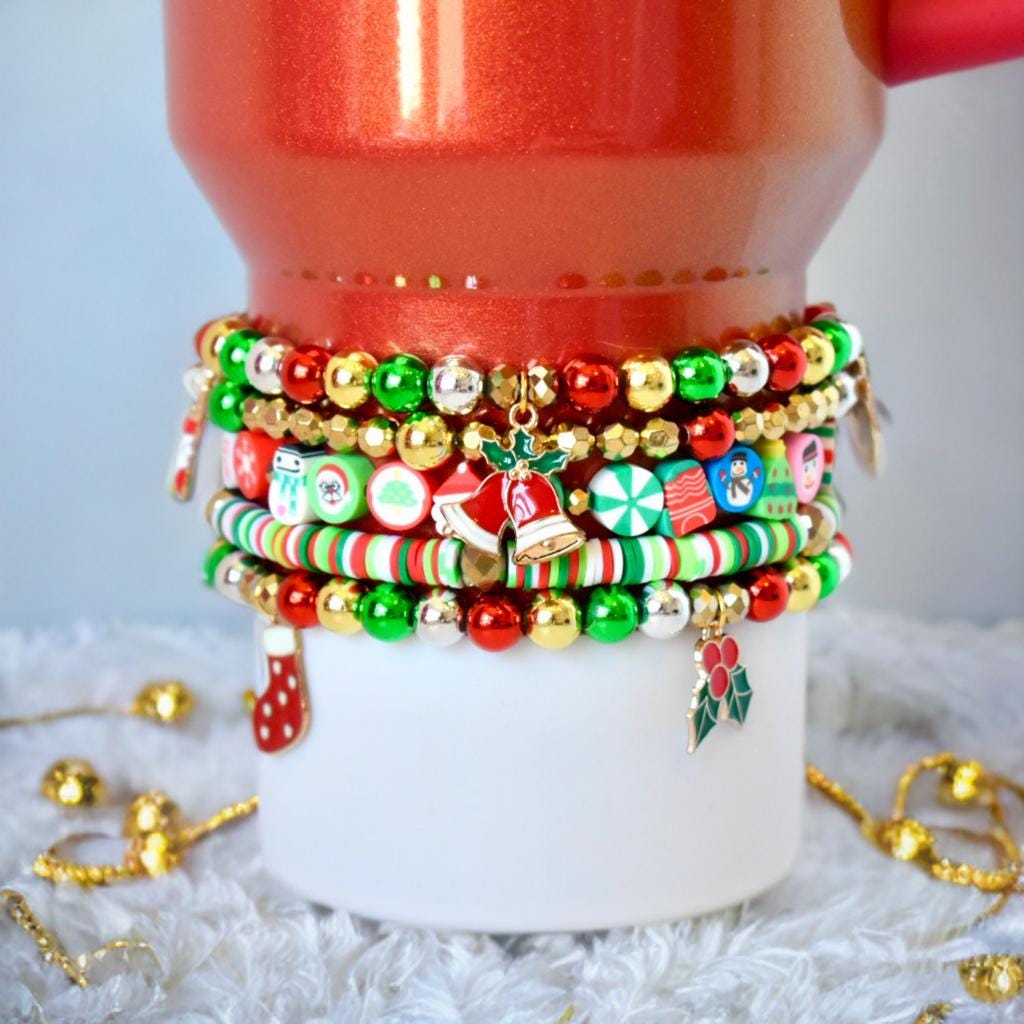 Christmas-themed beaded bracelets stacked on a white cylinder. The bracelets feature red, green, and gold beads, with charms including a bell, holly, and a stocking. The bracelets are festive, with candy cane and snowman-themed beads, perfect for holiday accessories.