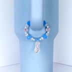 A light blue tumbler with a decorative handle charm. The charm features a circle of blue and pink beads, with a clear, iridescent slipper pendant. The tumbler has a handle and is set against a white background.