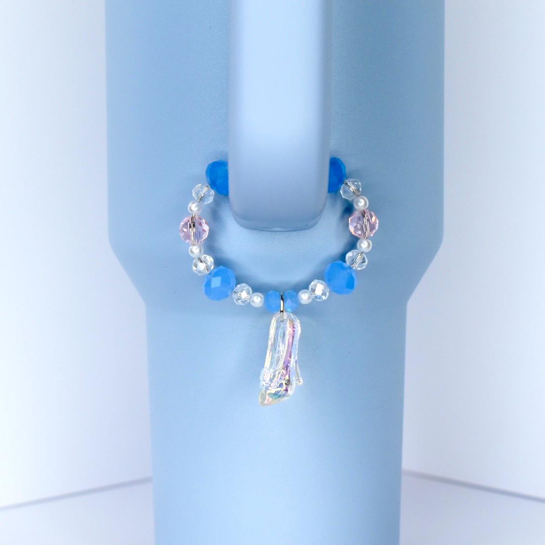 A light blue tumbler with a decorative handle charm. The charm features a circle of blue and pink beads, with a clear, iridescent slipper pendant. The tumbler has a handle and is set against a white background.