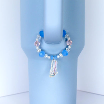 A light blue tumbler with a decorative handle charm. The charm features a circle of blue and pink beads, with a clear, iridescent slipper pendant. The tumbler has a handle and is set against a white background.