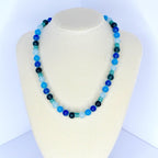 A beaded necklace with a variety of blue tones, from light sky blue to deep navy. The necklace is displayed on a white mannequin bust. The beads are round and evenly spaced, creating a simple yet elegant design.