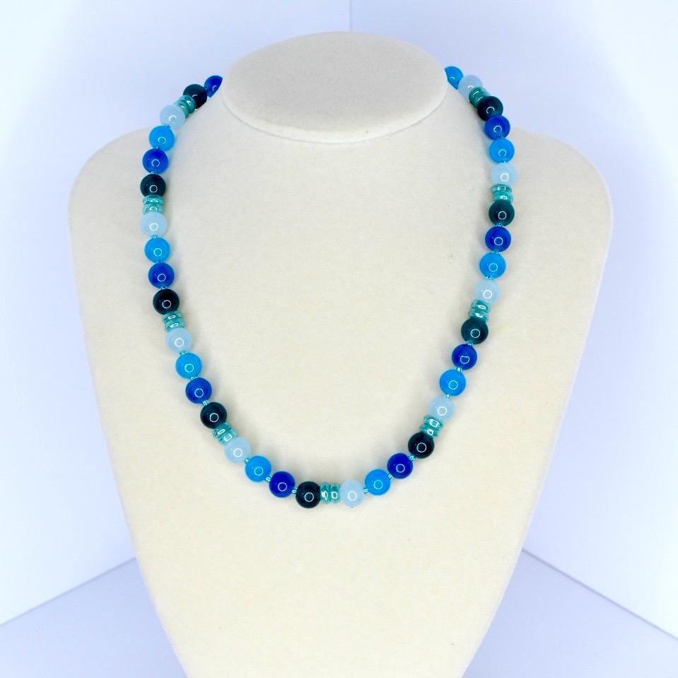 A beaded necklace with a variety of blue tones, from light sky blue to deep navy. The necklace is displayed on a white mannequin bust. The beads are round and evenly spaced, creating a simple yet elegant design.