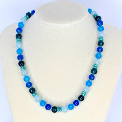 A beaded necklace with a gradient of blue hues, from light sky blue to deep navy. The necklace features round beads interspersed with smaller, cylindrical beads, creating a visually appealing accessory. The necklace is displayed on a white mannequin.